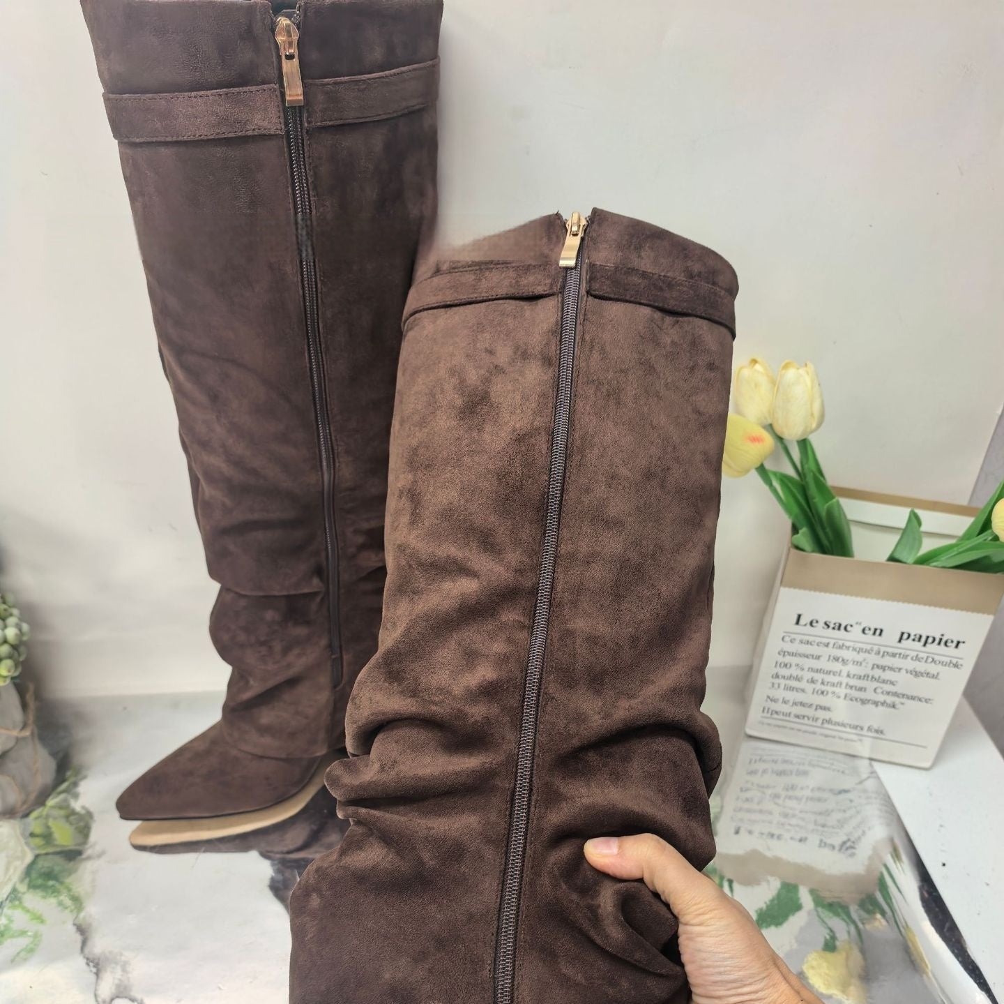 brown suede knee-high boots with zipper detail held by hand on reflective surface with flowers