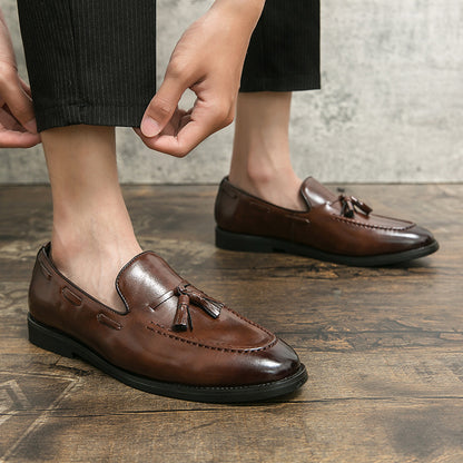 Men's brown tassel slip-on loafers in polished leather worn with black pants on wooden floor
