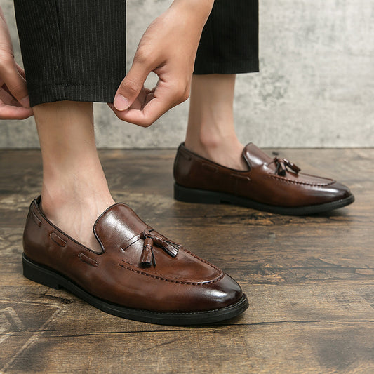 Men's brown tassel slip-on loafers in polished leather worn with black pants on wooden floor