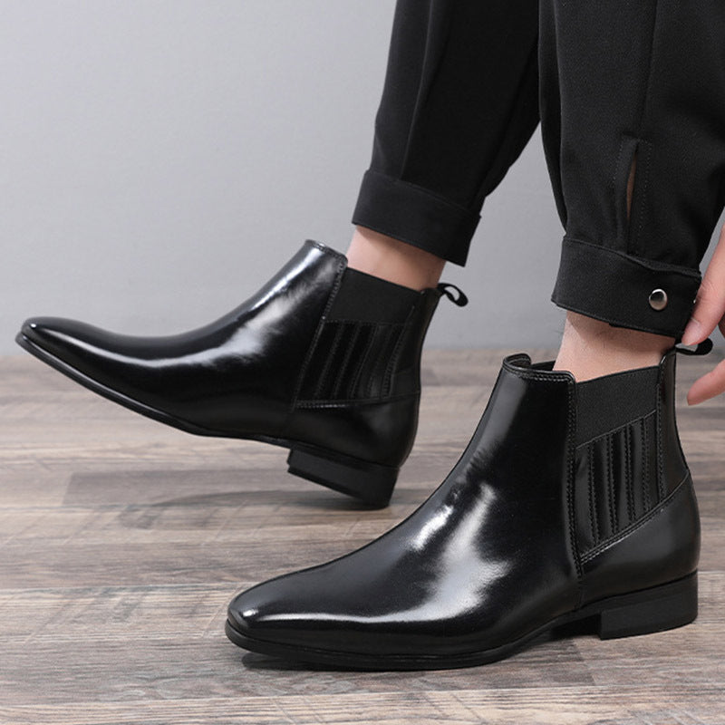 Men's black leather Chelsea boots with pointed toe and elastic side panels worn with black pants on wooden floor