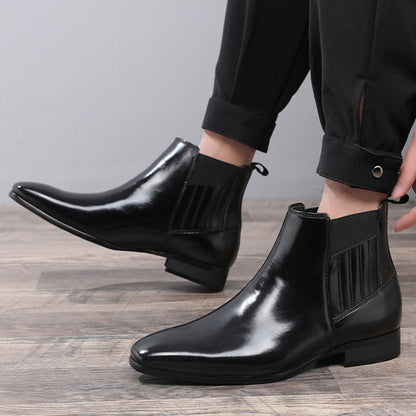 Men's black leather Chelsea boots with pointed toe and elastic side panels worn with black pants on wooden floor