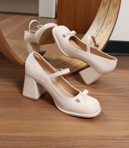 Beige patent leather Mary Jane shoes with chunky heels and bow detail on wooden floor in front of a round mirror