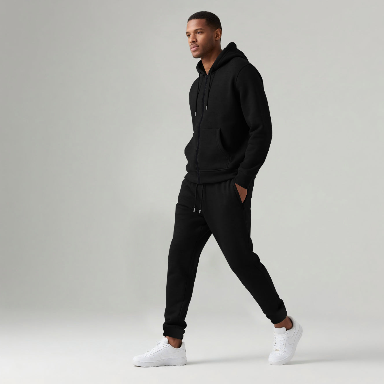 Man wearing black hooded zip-up sweatshirt and matching jogger pants with white sneakers on gray background