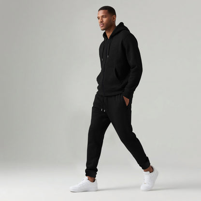 Man wearing black hooded zip-up sweatshirt and matching jogger pants with white sneakers on gray background
