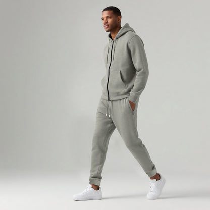 Man wearing light gray hooded zip-up sweatshirt and jogger sweatpants with white sneakers on gray background