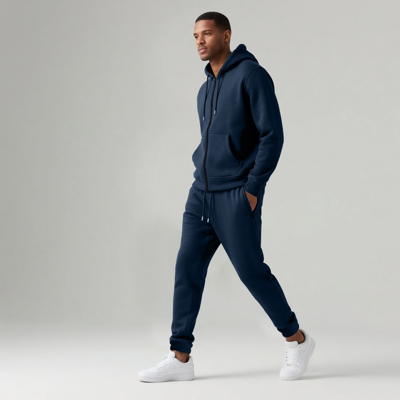 Man wearing navy blue hooded zip-up tracksuit and white sneakers walking on plain background