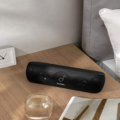 Black portable Bluetooth speaker with deep bass on wooden table next to glass and pillow