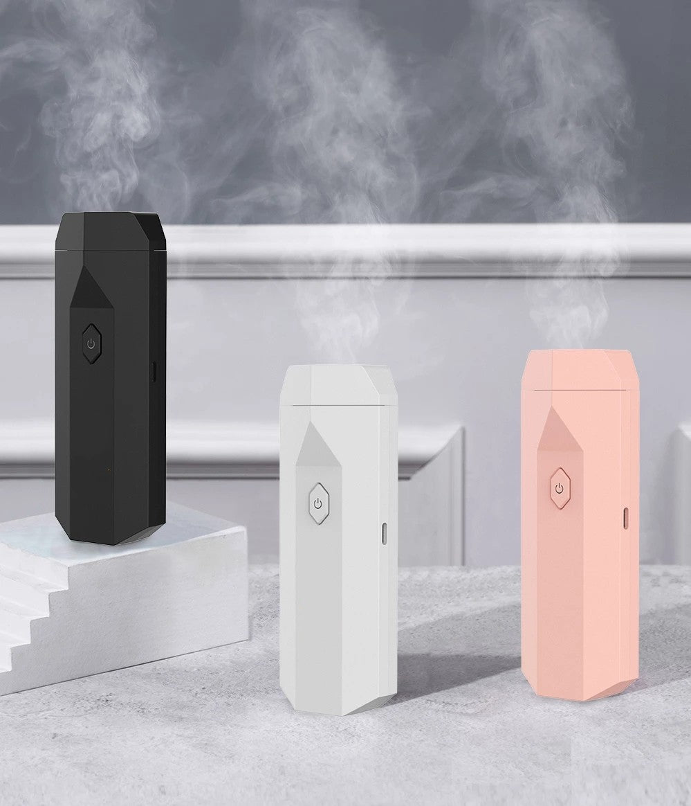 Black, white, and pink portable electronic bakhoor incense burners emitting smoke on a stone surface