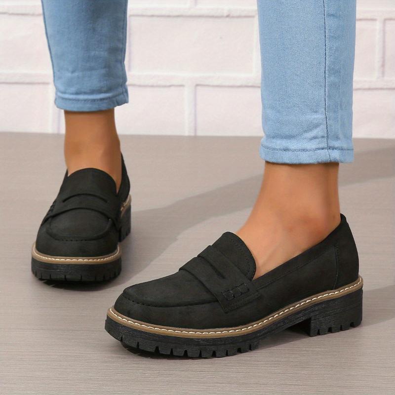Black thick-soled loafers worn with light blue rolled-up jeans on wooden floor