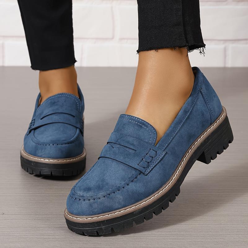 Close-up of blue suede women's loafers with thick black lug soles worn with black pants on a wooden floor
