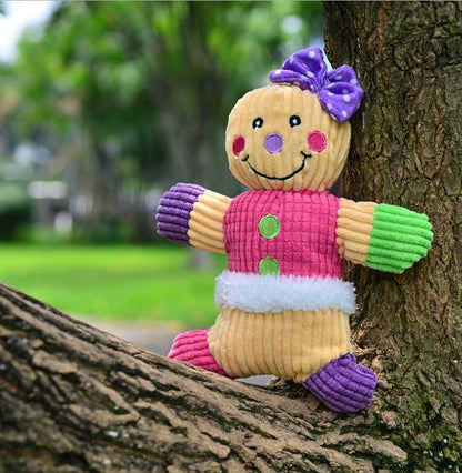 Colorful plush gingerbread man dog toy with purple bow, pink vest, and soft textured fabric on tree outdoors