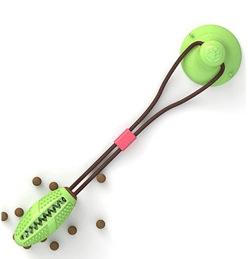 Green suction cup dog toy with brown elastic tug and textured chew end, surrounded by small brown kibbles