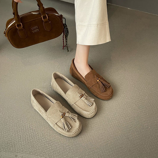 Women wearing brown suede tassel flat shoes next to beige suede tassel flats on gray floor
