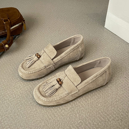 Beige women’s suede tassel flat loafers on carpeted floor next to brown suede handbag