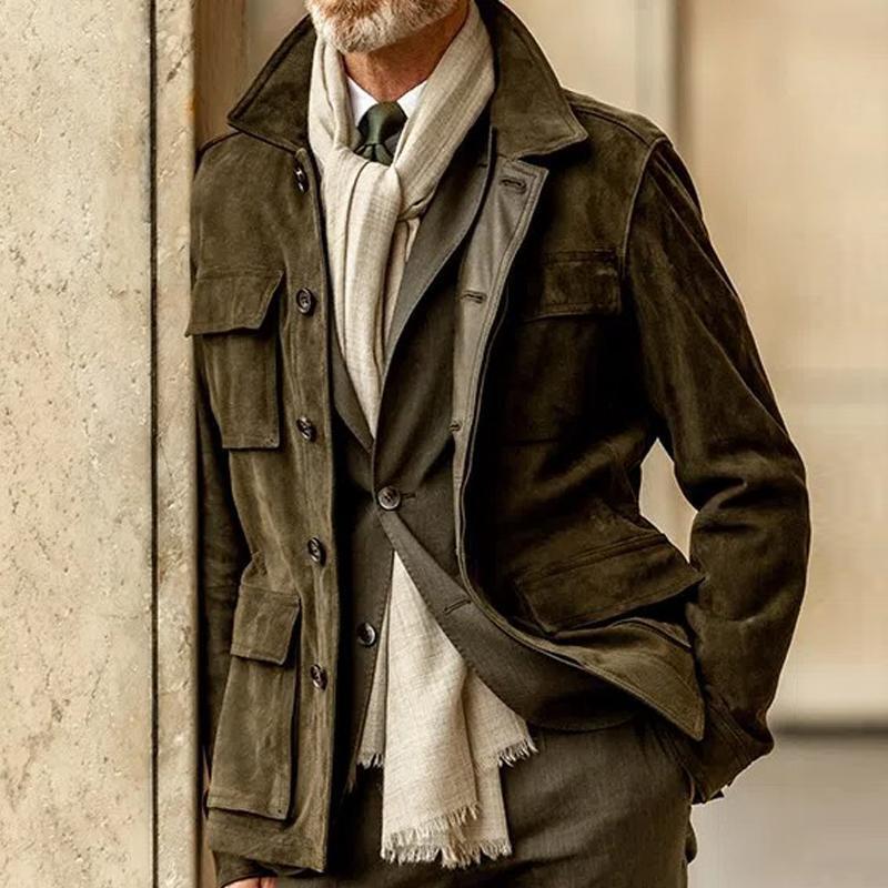 Man wearing brown suede jacket with flap pockets, beige scarf, and suit leaning on stone wall