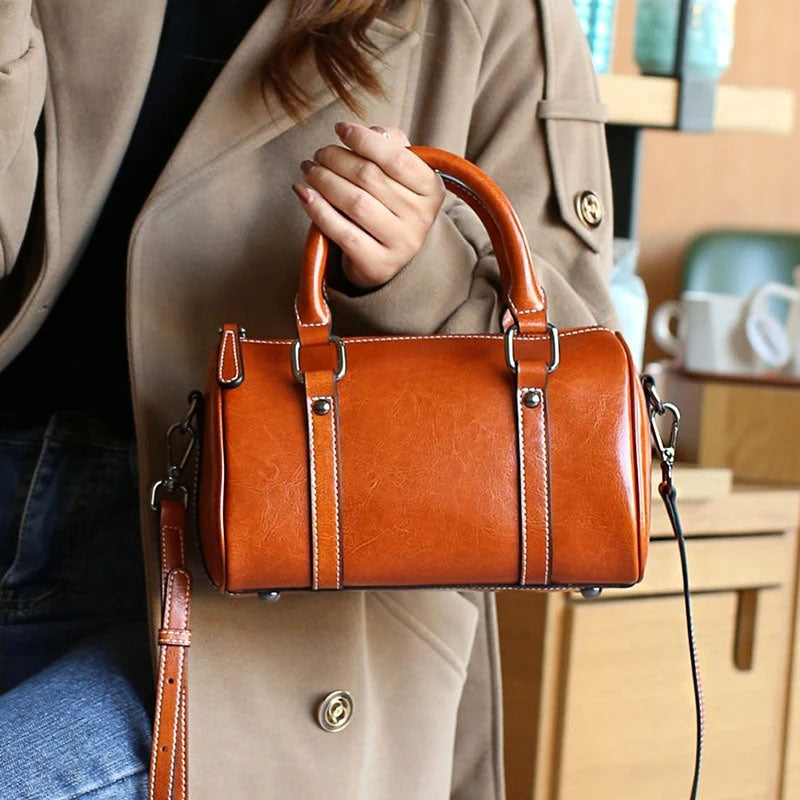 Caramel leather crossbody bag with double handles and silver hardware held by person in beige coat