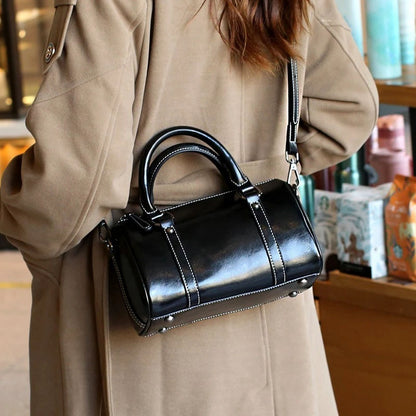 Black leather crossbody bag with white stitching worn over beige coat in indoor setting