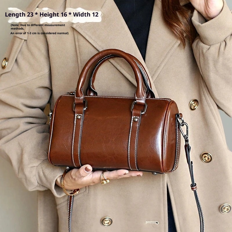 Caramel brown leather crossbody bag with dual handles and detachable strap, held by a woman in a beige coat