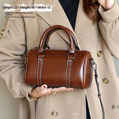 Caramel brown leather crossbody bag with dual handles and detachable strap, held by a woman in a beige coat