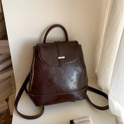 Brown PU leather small backpack with top handle and flap closure on white surface by beige wall