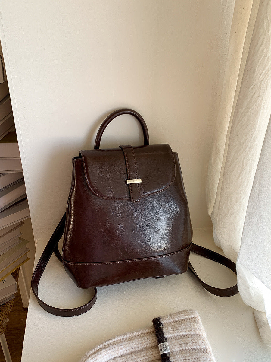 brown PU leather small backpack with top handle on white shelf near books and knitted accessory