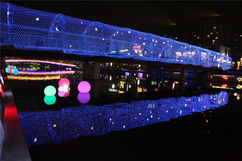Night scene of a pedestrian bridge adorned with blue LED net lights reflecting on calm water with colorful glowing spheres floating nearby