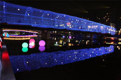 Night scene of a pedestrian bridge adorned with blue LED net lights reflecting on calm water with colorful glowing spheres floating nearby