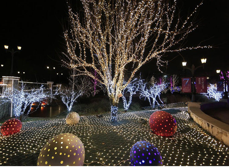 Outdoor night scene with leafless trees wrapped in warm white Christmas LED string lights and colorful illuminated decorative spheres