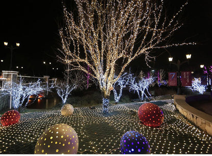 Outdoor night scene with leafless trees wrapped in warm white Christmas LED string lights and colorful illuminated decorative spheres