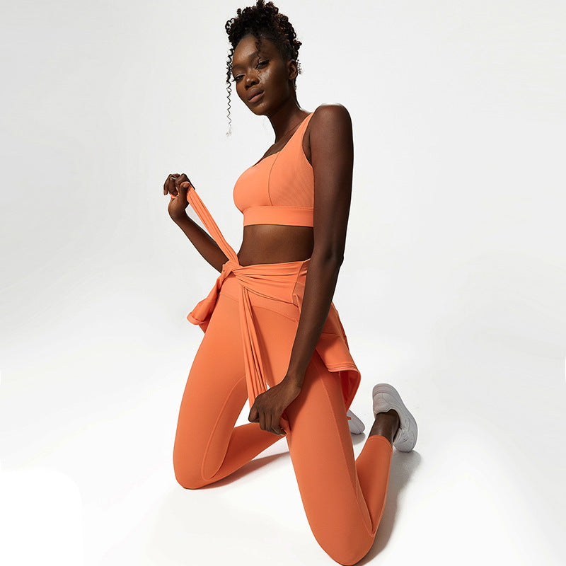 Woman in orange sports bra and leggings activewear set with sweatshirt tied around waist on white background