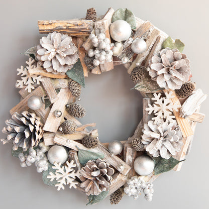 Silver and white Christmas wreath with pinecones, wooden snowflakes, silver baubles, and green leaves on gray background