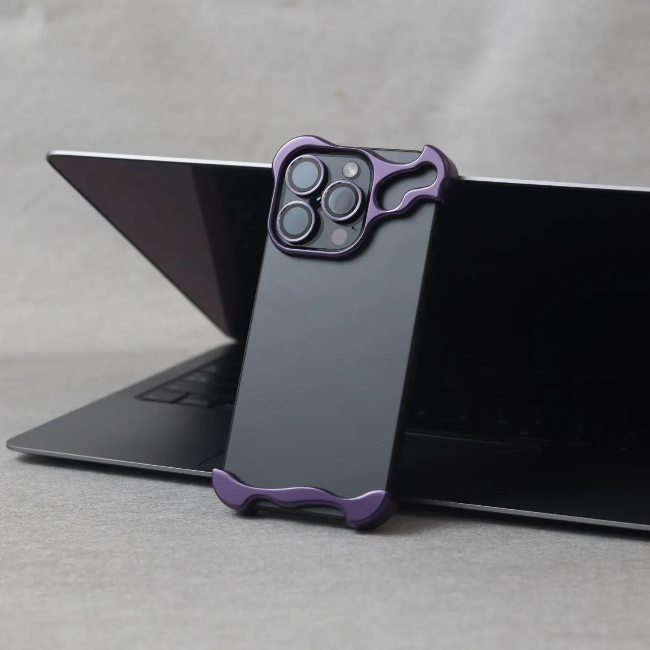 Black iPhone 15 Pro Max with purple zinc alloy protective case standing against a laptop
