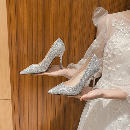Hands holding silver glitter pointed-toe bridal high heels with clear stiletto heels against wood panel background