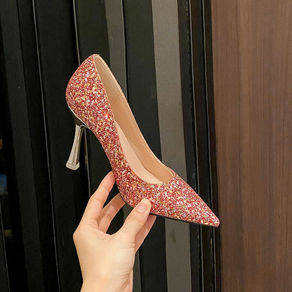Hand holding a red glittery pointed-toe high heel shoe with silver stiletto heel against a dark background