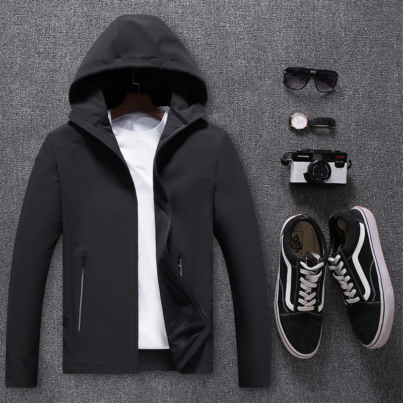 Black waterproof men's mountaineering jacket with hood, paired with white t-shirt, sunglasses, watch, camera, and black Vans sneakers on gray background