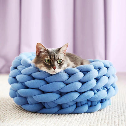 Gray tabby cat resting in a chunky knit blue round pet bed on a beige carpet with a lavender curtain backdrop