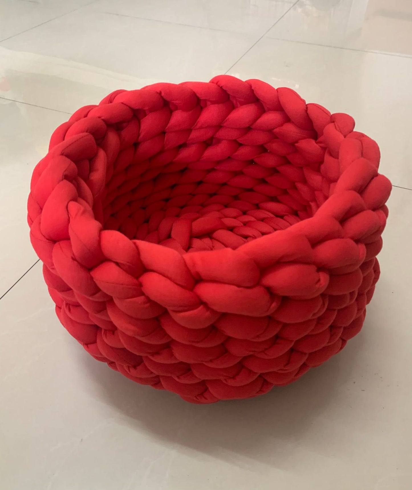 Red chunky knitted round pet bed on light tiled floor, soft cozy basket for small pets