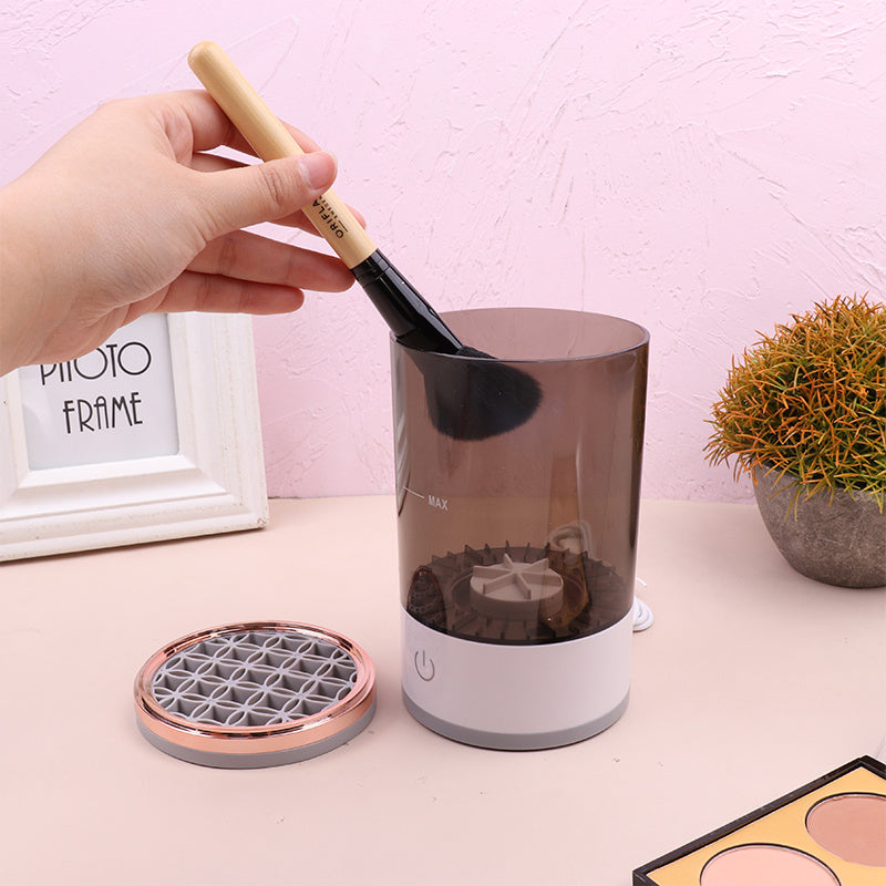 Portable electric makeup brush cleaner with wooden-handled brush being inserted in grey and white device on beige surface