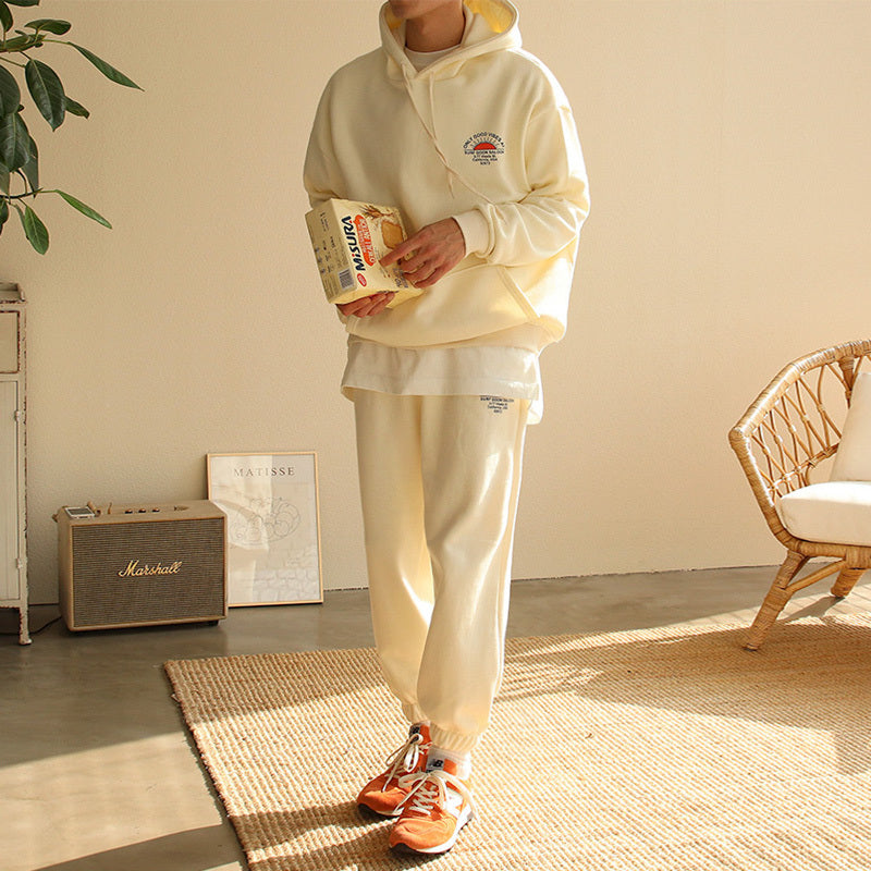Man wearing beige hooded sportswear set with orange sneakers holding a box in a cozy living room