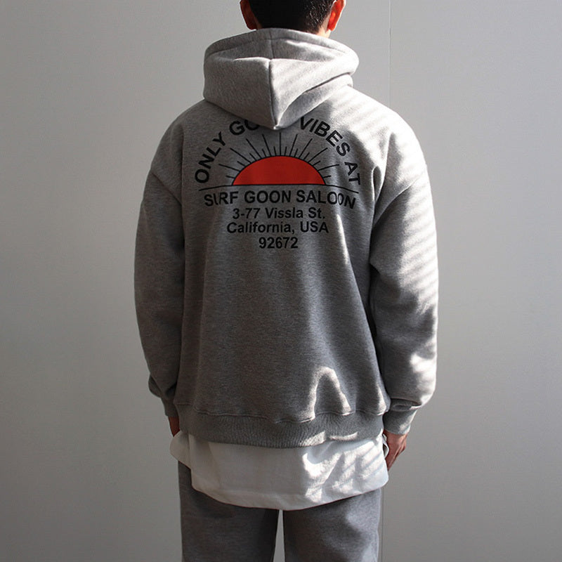 Man wearing grey hooded sweatshirt with red sun and black text graphic on back in casual indoor setting