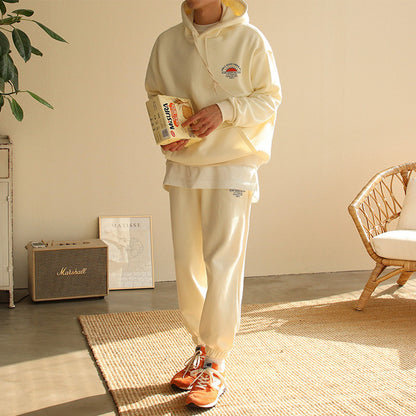 Man wearing cream hooded sweatshirt and joggers holding a cereal box, standing in a bright room with wicker chair and Marshall speaker