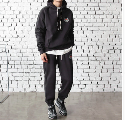 man wearing black hooded sportswear set with white drawstrings and sneakers against white brick wall