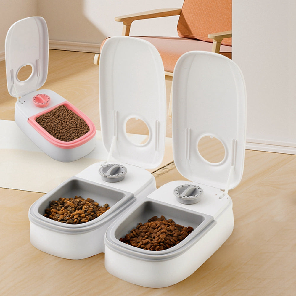 Three automatic pet feeders with open lids, filled with dry food, on a wooden floor with a chair in background