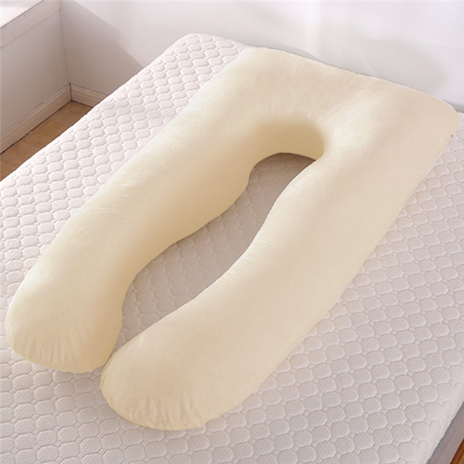 Beige U-shape maternity pillow on a white quilted mattress in a bright room