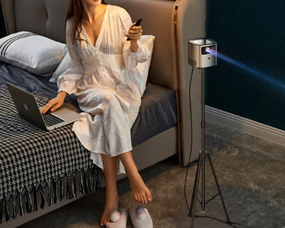 Woman in white dress sitting on bed using laptop and remote with portable projector on tripod in dark room