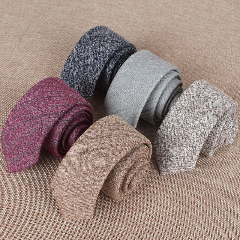 Five slim cotton ties in burgundy, navy, gray, beige, and light brown rolled on a textured brown fabric background