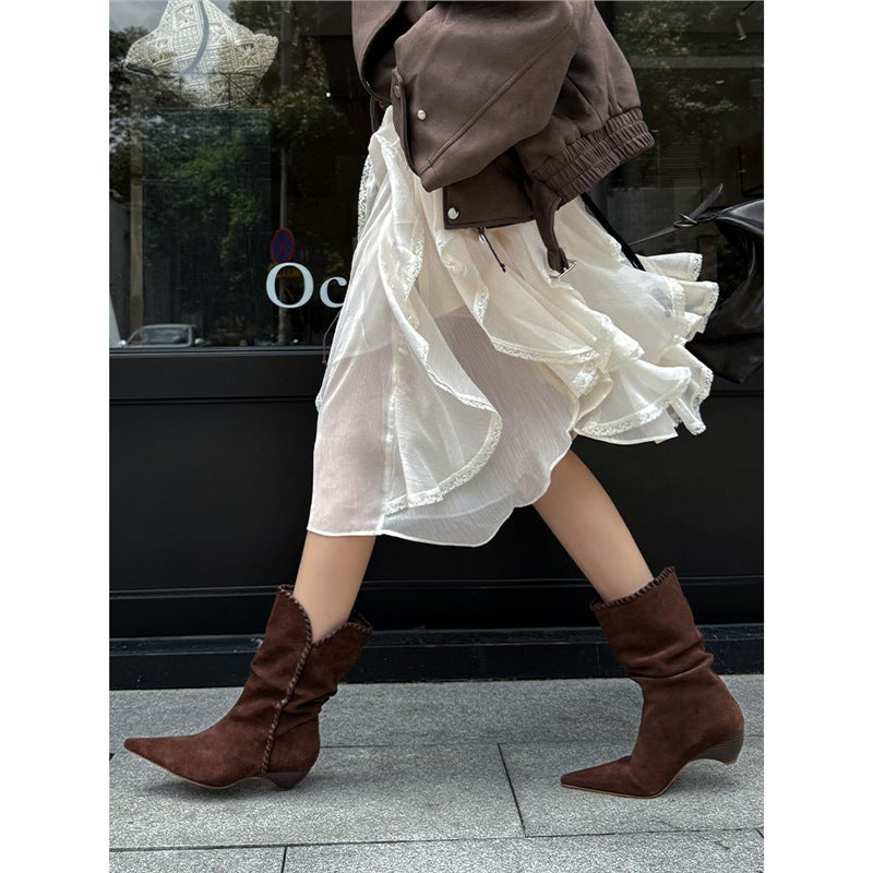 Brown suede pointed toe Western boots worn with flowing white skirt and brown jacket on city sidewalk
