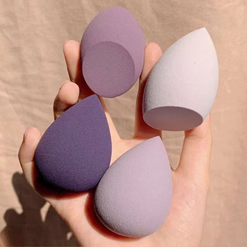 Hand holding four teardrop-shaped makeup sponges in shades of purple and nude for blending foundation