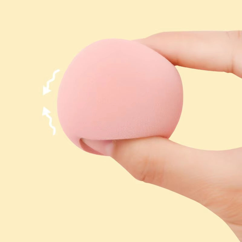 Close-up of a hand gently squeezing a soft, round pink makeup sponge against a pale yellow background