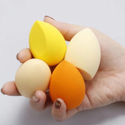 Hand holding four colorful makeup sponges in yellow, beige, orange, and cream tones
